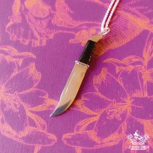 K&ouml;nnte beinhalten: Eine silberne Messer-Anh&auml;nger-Kette mit schwarzem Griff. Das Messer h&auml;ngt an einer silbernen Kette vor einem violetten Hintergrund mit orangefarbenen Blumendesigns.