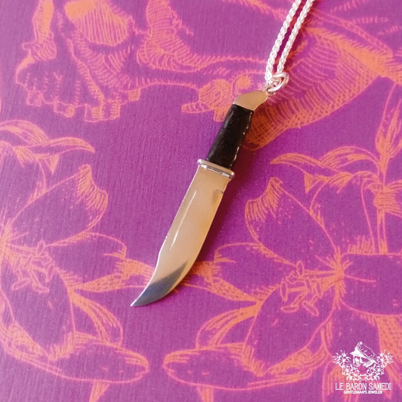 K&ouml;nnte beinhalten: Eine silberne Messer-Anh&auml;nger-Kette mit schwarzem Griff. Das Messer h&auml;ngt an einer silbernen Kette vor einem violetten Hintergrund mit orangefarbenen Blumendesigns.