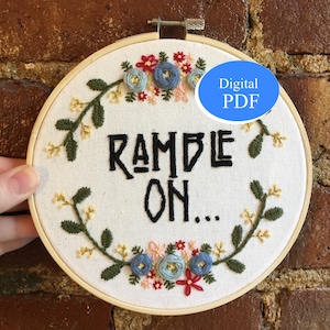 May include: Embroidered hoop art with a floral design and the text "RAMBLE ON..." in black lettering. The hoop is made of wood and has a white fabric background. The flowers are embroidered in shades of blue, red, and pink.