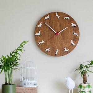 May include: A brown wooden clock with white dog silhouettes around the face. The clock hands are black and red. The clock is hanging on a white wall.