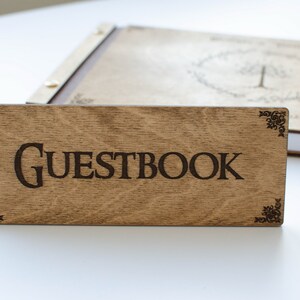Pu&ograve; includere: Un cartello in legno con la parola "GUESTBOOK" incisa in lettere nere. Il cartello ha angoli decorativi ed &egrave; appoggiato su una superficie bianca.