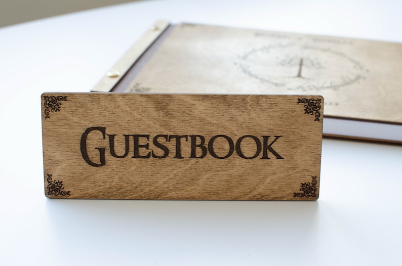 Pu&ograve; includere: Un cartello in legno con la parola "GUESTBOOK" incisa in lettere nere. Il cartello ha angoli decorativi ed &egrave; appoggiato su una superficie bianca.