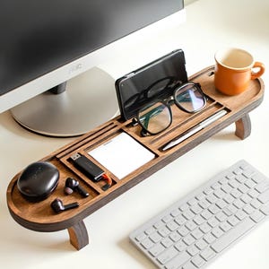 May include: A wooden desk organizer with multiple compartments holds a smartphone, glasses, pen, notepad, and USB drive. A black wireless earbud case and earbuds are also present. An orange mug sits on the right side. The organizer is on a white desk.