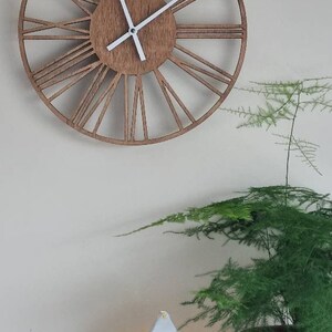 May include: A white metal lantern with a clear glass panel and a string of fairy lights inside. The lantern has a star design and is sitting on a white surface in front of a brown wooden clock with Roman numerals.