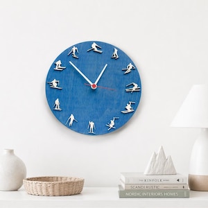 Può includere: Un orologio da parete blu con lancette bianche e lancetta dei secondi rossa. Il quadrante presenta piccole figure di sciatori bianchi al posto dei numeri. L'orologio è montato su una parete bianca.