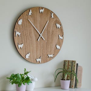 Può includere: Un orologio da parete rotondo in legno con finitura marrone chiaro. Il quadrante presenta sagome di gatti bianchi che segnano le ore. Le lancette dell'orologio sono bianche. L'orologio è montato su una parete bianca.