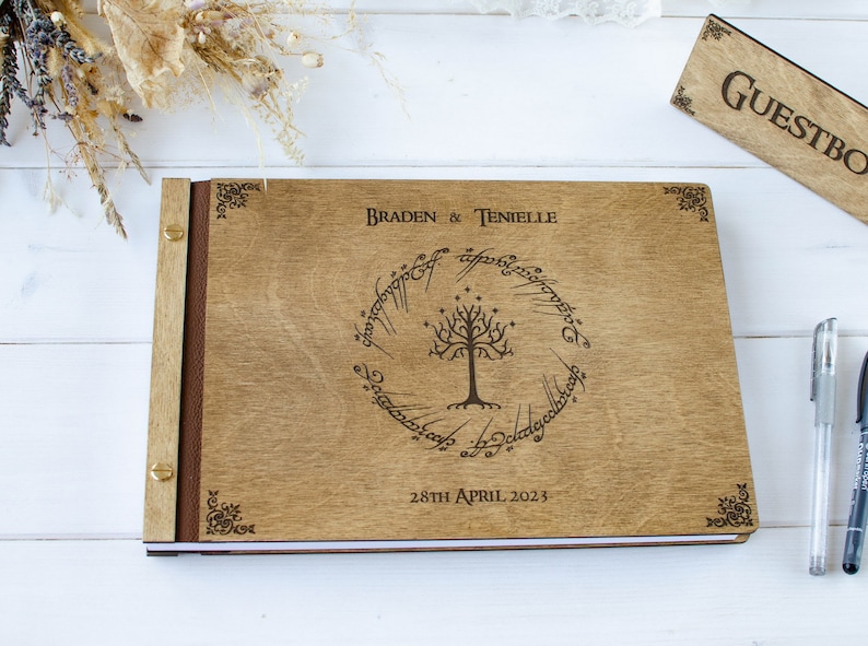 Pu&ograve; includere: Un libro degli ospiti in legno con una spina dorsale in pelle marrone e accenti in metallo dorato. La copertina anteriore presenta un design inciso a laser dell'Albero Bianco di Gondor dal Signore degli Anelli, circondato dall'iscrizione "Un anello per domarli tutti, Un anello per trovarli, Un anello per portarli tutti e nell'oscurit&agrave; legarli." I testi "Braden & Tenielle" e "28th April 2023" sono anche incisi sulla copertina.