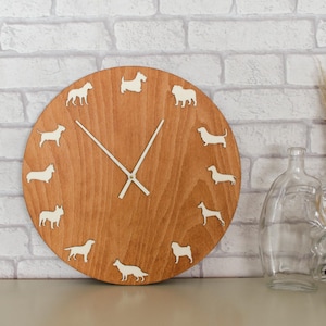 Puede incluir: Un reloj de madera redondo con acabado marrón claro. La esfera del reloj presenta siluetas de perros blancos que marcan las horas. Las manecillas del reloj son blancas. El fondo es una pared de ladrillo blanco. Botellas de vidrio y una figura de pájaro negro sobre la mesa.