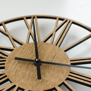 A brown wooden clock with black Roman numeral hour markers and black hands. The clock has a geometric design with black lines radiating from the center.