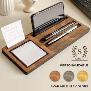 Puede incluir: Organizador de escritorio de madera con ranuras para teléfono, bolígrafos y bloc de notas. El texto "STARVE YOUR DISTRACTIONS, FEED YOUR FOCUS" y "SIMON PERSONALIZABLE" son visibles. Disponible en tres colores.