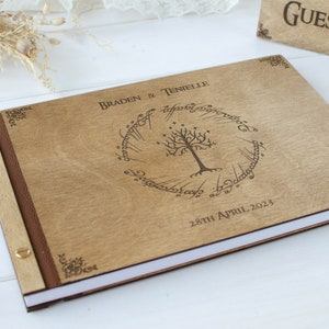 Pu&ograve; includere: Un libro degli ospiti in legno con una copertina marrone e un design inciso a laser di un albero con un anello attorno. Il testo "Braden & Tenielle" e "28th April 2023" &egrave; inciso sulla copertina.