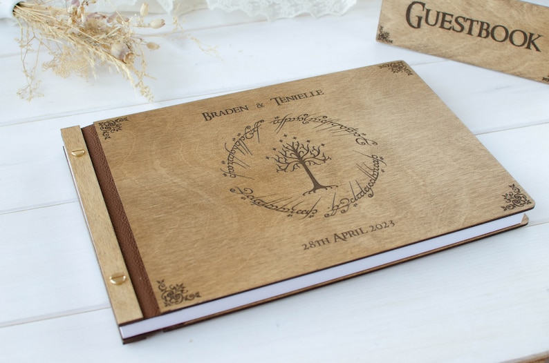 Pu&ograve; includere: Un libro degli ospiti in legno con una copertina marrone e un design inciso a laser di un albero con un anello attorno. Il testo "Braden & Tenielle" e "28th April 2023" &egrave; inciso sulla copertina.