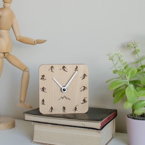Könnte beinhalten: Eine quadratische Holzuhr mit hellem Holzfinish. Das Zifferblatt zeigt schwarze Skifahrer-Silhouetten und eine Bergkette. Weiße Zeiger zeigen die Uhrzeit an. Die Uhr steht auf zwei Büchern. Ungefähre Abmessungen: 15 cm x 15 cm.