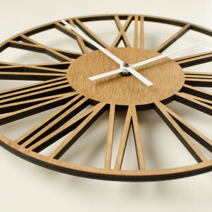 May include: A large, round, wooden wall clock with Roman numeral markings and white hands. The clock face is made of wood with a light brown finish. The clock has a decorative, open-work design.