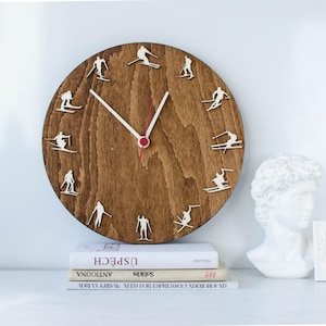 May include: A round wooden wall clock with a dark brown finish. The clock features white skier silhouettes around the perimeter and white hands with a red center. The clock is approximately 12 inches in diameter.