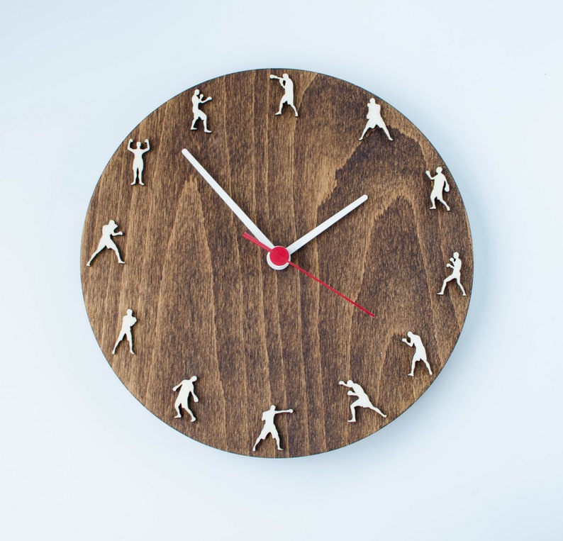 Puede incluir: Un reloj de madera marr&oacute;n con agujas blancas y una aguja de segundos roja. La esfera del reloj tiene una silueta de un boxeador en cada posici&oacute;n de hora.
