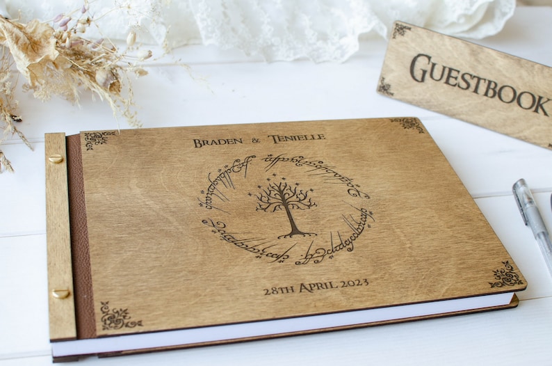 Pu&ograve; includere: Un libro degli ospiti in legno con una spina dorsale in pelle marrone. La copertina presenta un design inciso dell'Albero Bianco di Gondor dal Signore degli Anelli, circondato dall'iscrizione "Un solo anello per governarli tutti." Il libro &egrave; intitolato "Braden & Tenielle" e la data "28 aprile 2023" &egrave; stampata sotto il design.