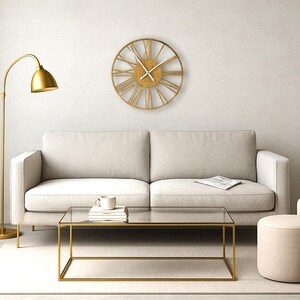 May include: A gold-toned metal clock with Roman numeral markings hangs on a white wall above a white sofa. A gold floor lamp, a glass coffee table, and two beige ottomans are in the room.