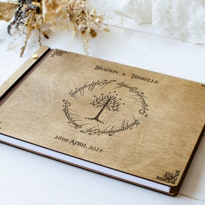 Pu&ograve; includere: Un libro degli ospiti in legno con una rilegatura in pelle marrone e un design inciso a laser che presenta l'Unico Anello de Il Signore degli Anelli, un albero e l'iscrizione "Braden & Tenielle 28 aprile 2023".