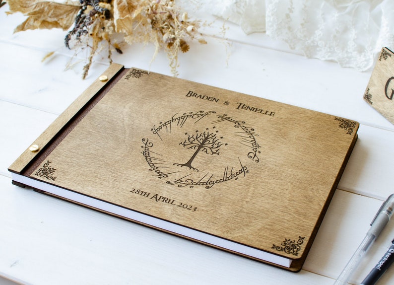 Pu&ograve; includere: Un libro degli ospiti in legno con una rilegatura in pelle marrone e un design inciso a laser che presenta l'Unico Anello de Il Signore degli Anelli, un albero e l'iscrizione "Braden & Tenielle 28 aprile 2023".