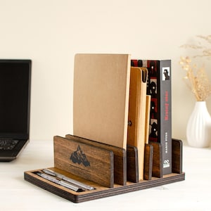 May include: Wooden desk organiser with a mountain logo, holding notebooks, folders, and a book. Two pens rest in the front tray. The organiser is made of dark wood and sits on a white surface.