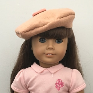 May include: A doll wearing a pink polo shirt with a pink heart design, a brown beret with a pink button, and a plaid skirt.