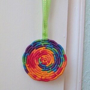 Op de afbeelding: Een rond, geweven ornament met een spiraalontwerp in levendige regenboogkleuren: rood, oranje, geel, groen, blauw en paars. Een groen lint is bevestigd om op te hangen. Het ornament is waarschijnlijk handgemaakt en decoratief.