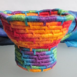 Könnte beinhalten: Ein handgewebter Korb in Form einer Teetasse mit einem lebendigen Regenbogen an Farben. Der Korb hat einen Henkel und besteht aus gewebtem Garn. Die Farben wechseln von Lila zu Blau, Grün, Gelb, Orange und Rot.