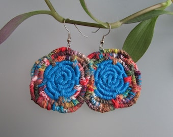 May include: A pair of colorful, round earrings with a blue center. The earrings are made of woven fabric and have a silver hook.