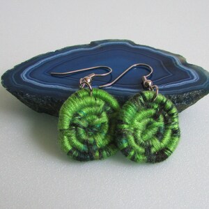 May include: A pair of green and black crocheted earrings with silver hooks. The earrings are round and have a spiral design.