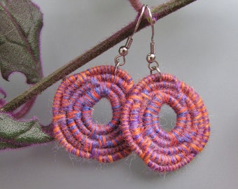 May include: A pair of purple and orange circular earrings with a woven design. The earrings are made of yarn and have silver hooks.