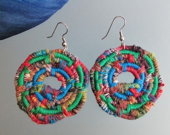 May include: A pair of colorful, circular earrings made from woven fabric. The earrings are a mix of green, blue, red, brown, and white fabric. The earrings are attached to silver hooks.