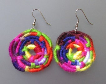 May include: A pair of colorful crocheted earrings with a circular design. The earrings are made with a variety of colors, including pink, purple, blue, green, yellow, and orange. The earrings are attached to silver hooks.