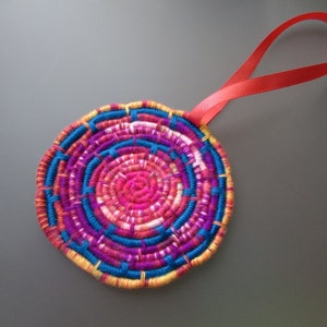Może przedstawiać: Kolorowy, okrągły, tkany ornament z czerwoną wstążką do zawieszenia. Ornament jest wykonany z przędzy w odcieniach różowego, niebieskiego, żółtego i białego.