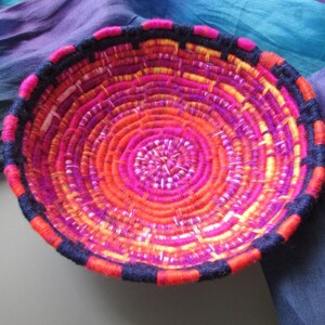 Può includere: Un cesto intrecciato di forma circolare con un design colorato. Il cesto è fatto di filo rosa, arancione e giallo, con un bordo nero.