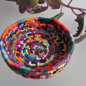 May include: A colorful woven basket made from yarn in a variety of colors, including red, orange, yellow, green, blue, purple, and white. The basket is round and has a circular opening. The basket is hanging from a plant.