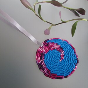 Peut inclure: Une décoration ronde en tissage bleu et rose avec un ruban argenté attaché. La décoration a un motif en spirale au centre.
