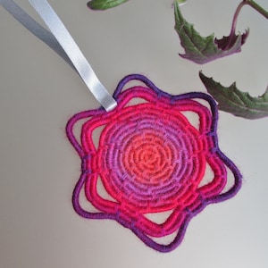 Puede incluir: Un tapiz de macramé en forma de estrella de color rosa y morado degradado con una cinta plateada.