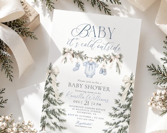 Könnte beinhalten: Eine weiße Babyparty-Einladungskarte mit dem Text Baby it's cold outside, einem blauen Body, Socken und Babyschuhen an einer Tannengirlande sowie zwei schneebedeckten Tannenbäumen mit cremefarbenen Schleifen an den Seiten.