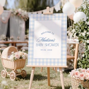 Blue gingham baby shower welcome sign template displayed on an easel at a soft outdoor baby shower with florals, balloons and elegant pastel decor.