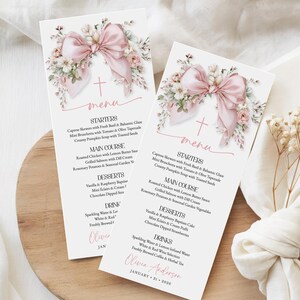 May include: Two elegant menu cards with a soft pink bow and floral design. The cards feature a cross and the word "menu" in a delicate script. The menu includes starters, main courses, desserts, and drinks, perfect for a baptism or wedding.