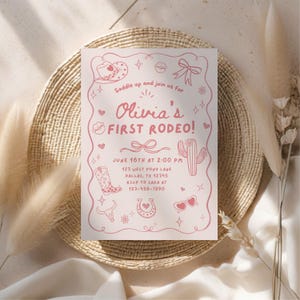 May include: A pink invitation card with the text "Olivia's First Rodeo!" and rodeo-themed illustrations. The card includes event details such as date, time, and location. The card is placed on a woven, circular placemat.