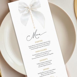 May include: A white menu card with a decorative bow and rosary bead design. The menu lists starters, main courses, desserts, and drinks. The menu is on a white plate.