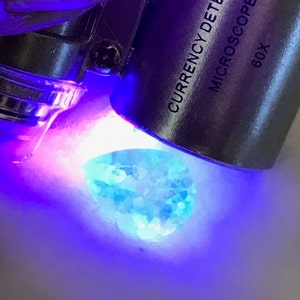 May include: A blue gemstone is illuminated by a UV light from a currency detector microscope labeled "CURRENCY DETECTOR MICROSCOPE 60X".