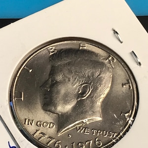 Puede incluir: Una moneda de medio dólar de plata de 1976, con el perfil de un hombre y las inscripciones "IN GOD WE TRUST" y "LIBERTY". La moneda está dentro de un soporte de cartón blanco, asegurado con grapas metálicas.