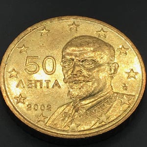 Puede incluir: Moneda dorada con el número "50" y la palabra "ЛEПTA". La moneda presenta un retrato de un hombre con gafas y bigote, rodeado de estrellas. También se muestra el año "2002".
