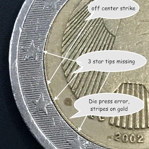 Puede incluir: Primer plano de una moneda de dos euros de 2002 con errores visibles. La moneda muestra un golpe descentrado, tres puntas de estrella faltantes y errores de prensa con rayas en la sección dorada. El borde tiene un diseño plateado y acanalado.