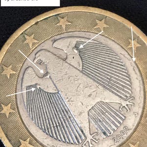 Puede incluir: Una moneda de euro alemana de 2002 con un águila y flechas blancas que señalan tres gotas de metal, una acuñación descentrada y un cuño agrietado. Un recuadro blanco contiene el texto: 1) 3 metal run blobs, 2) off center strike, 3) cracked die.