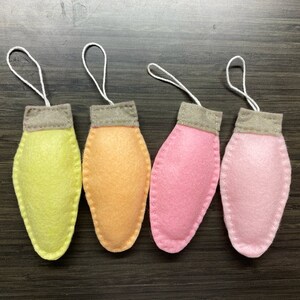 May include: Four felt Christmas light ornaments in pastel colors: yellow, orange, pink, and light pink. Each ornament has a gray cap and a white string for hanging. The ornaments are handmade and suitable for holiday decor.