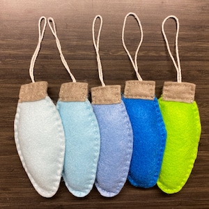 May include: Five handmade felt Christmas light ornaments in shades of blue and green, with tan tops and white string loops for hanging. The ornaments are arranged in a row against a dark wood grain background.